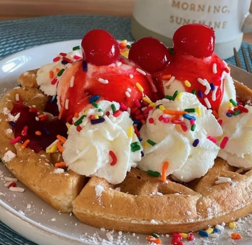 A delicious waffle topped with whipped cream, cherries, ice cream, and colorful sprinkles, served on a plate.