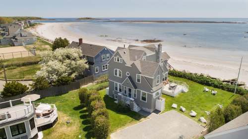 Aerial view of a coastal house with a lawn, beach, and water in the background on a sunny day.