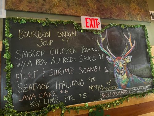 Chalkboard menu featuring dishes like Bourbon Onion Soup, Smoked Chicken Ravioli, and a colorful deer illustration.