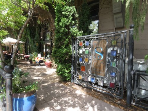 A decorative gate with a large "S" surrounded by colorful tiles, leading to a lush garden area with seating.