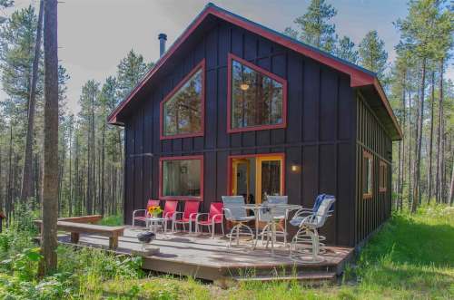 A modern black cabin with red accents, surrounded by tall trees and featuring a deck with outdoor seating.