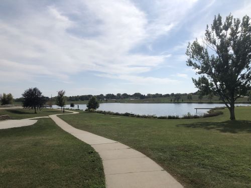 A winding path leads to a serene lake surrounded by trees and grassy areas under a partly cloudy sky.