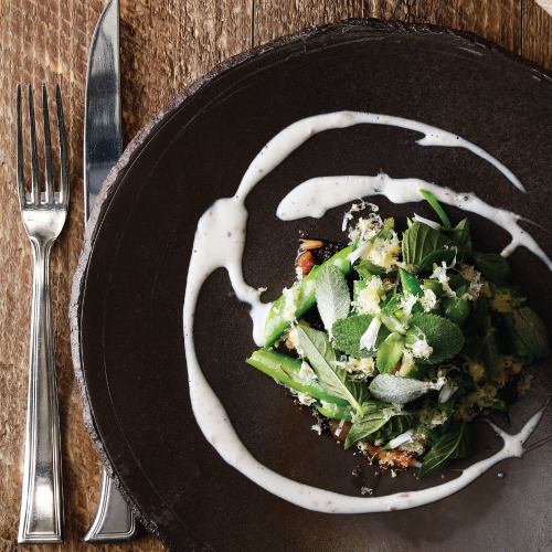 A gourmet dish featuring green vegetables and herbs, drizzled with a creamy sauce, served on a dark plate.