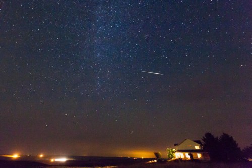 A starry night sky with a bright meteor streaking across, above a house illuminated by warm lights.