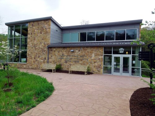 A modern stone and glass building with benches outside, surrounded by greenery and a paved walkway.