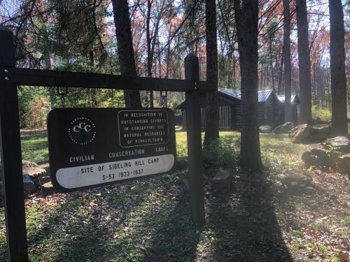 Sign for Sideling Hill Camp, a Civilian Conservation Corps site, surrounded by trees and rustic buildings.