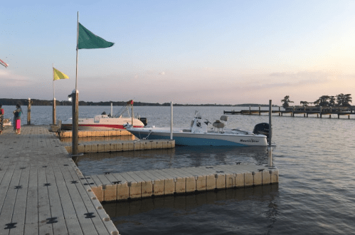 A peaceful waterfront scene with boats docked at a wooden pier, flags waving in the breeze under a pastel sky.
