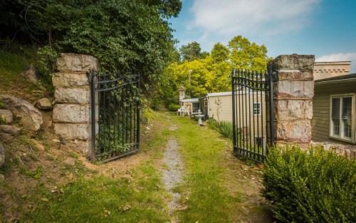 A stone gate opens to a grassy path leading to a house, surrounded by trees and greenery under a blue sky.