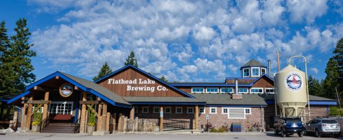 Exterior of Flathead Lake Brewing Co. featuring a rustic building, blue accents, and a large brewing tank.