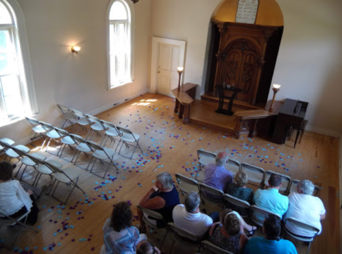 A small gathering in a light-filled room with chairs arranged facing a wooden stage and scattered blue items on the floor.