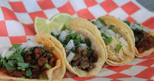 Four tacos on a checkered paper plate, topped with onions and cilantro, with a lime wedge on the side.
