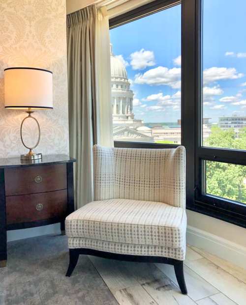 A cozy chair beside a lamp, with a view of a dome building and blue sky through a large window.