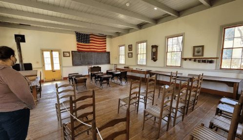 A historic classroom with wooden desks, chairs, and an American flag on the wall, featuring large windows and a chalkboard.