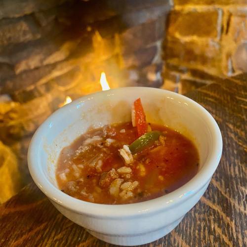 A bowl of hearty chili with meat, rice, and vegetables, set against a warm, glowing fireplace.