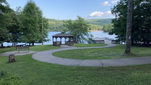 A serene lakeside scene featuring a gazebo, winding path, and lush greenery under a clear blue sky.