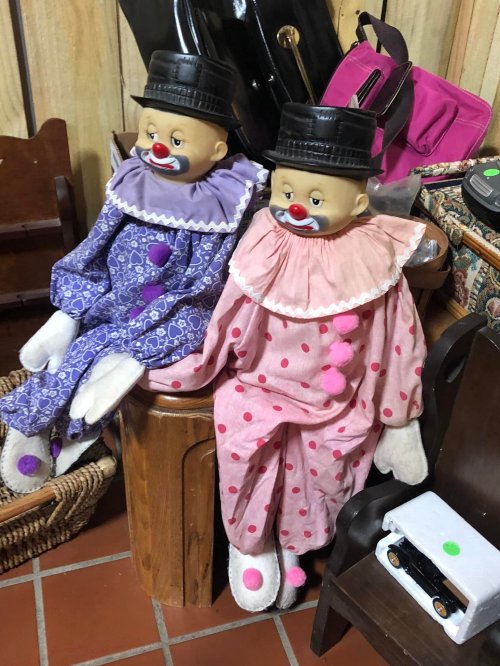 Two vintage clown dolls in colorful outfits, sitting on a wooden stool among various bags and items.