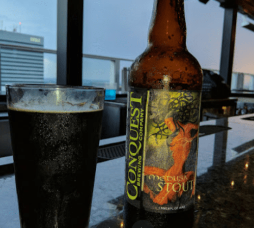 A bottle of Conquest Brewing Company's Medusa Stout next to a glass of dark beer on a bar counter.