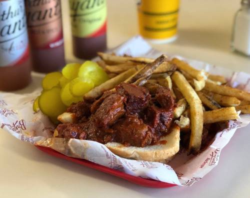 A plate of barbecue beef on bread, served with fries and pickles, with drinks in the background.