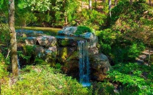 A serene waterfall cascades over rocks, surrounded by lush greenery and trees in a peaceful natural setting.