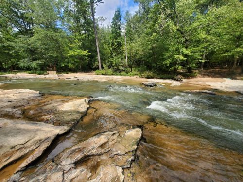 A serene river flows over rocky terrain, surrounded by lush green trees under a partly cloudy sky.