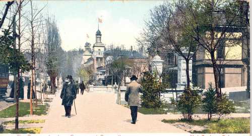 A vintage postcard showing a tree-lined street with people walking, historic buildings, and a flag atop a tower.