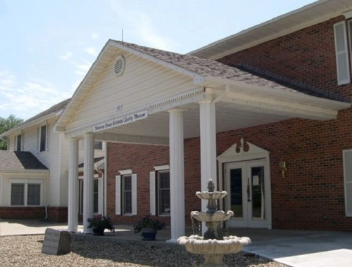 Exterior view of a brick building with a white porch and columns, featuring a fountain in front.