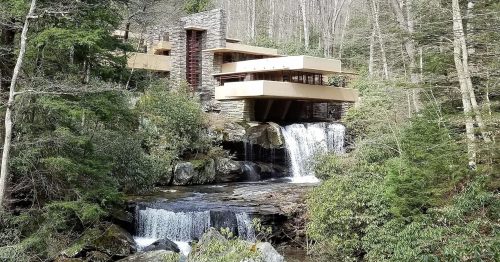 A modern house built over a waterfall, surrounded by lush greenery and rocky terrain.