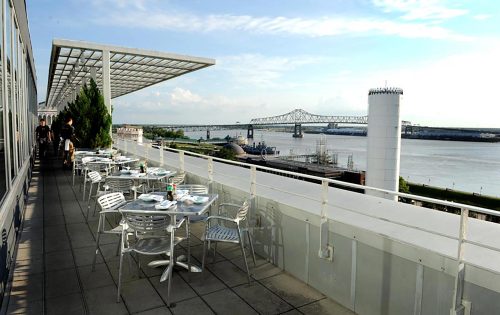 A rooftop terrace with tables overlooking a river and bridge, under a clear blue sky.