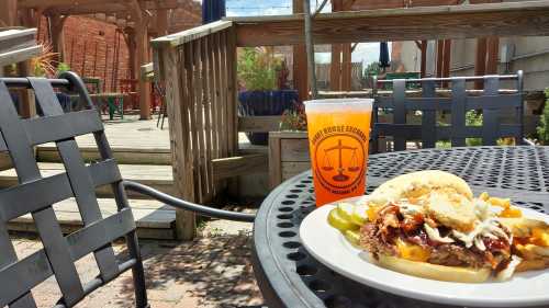 A burger with fries and pickles on a table, with a drink beside it, set in a sunny outdoor dining area.