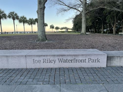 Stone bench with the inscription "Joe Riley Waterfront Park" in a park setting with palm trees and a clear sky.