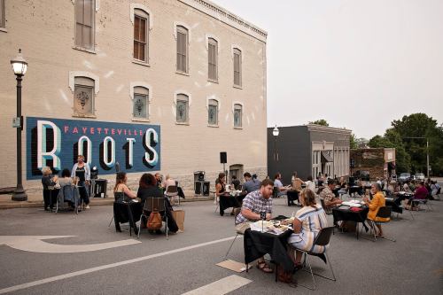 Outdoor dining scene with people at tables in front of a mural reading "Fayetteville ROOTS" on a city street.