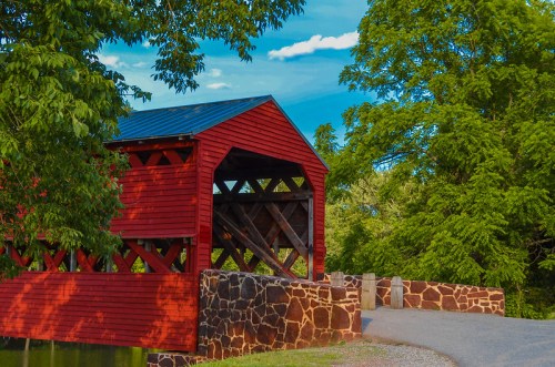 A red covered bridge stands over a stone path, surrounded by lush green trees and a clear blue sky.