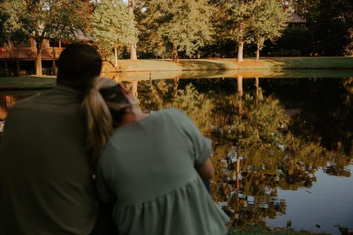 A couple sits by a serene pond, surrounded by trees, enjoying a peaceful moment together.