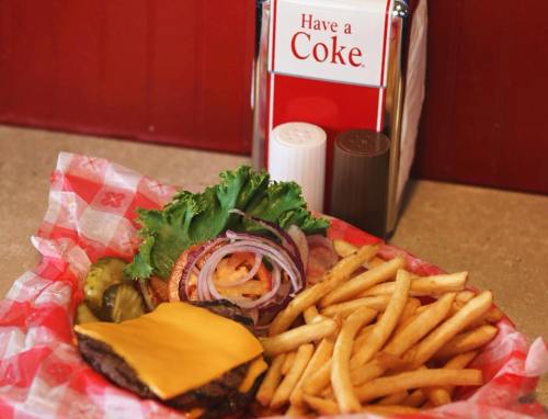 A cheeseburger with lettuce, onions, pickles, and fries in a basket, with a "Have a Coke" sign in the background.
