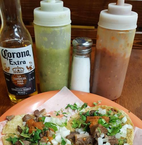 A plate of tacos topped with cilantro, alongside bottles of sauces, a salt shaker, and a Corona beer.