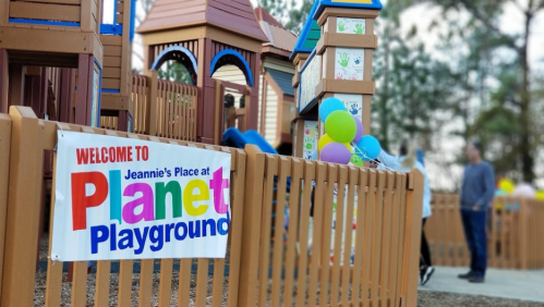 Colorful sign reading "Welcome to Jeannie's Place at Planet Playground" near a playground with balloons and people.