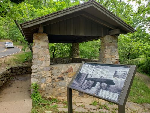 A stone shelter with a roof, surrounded by greenery, featuring an informational sign in front.