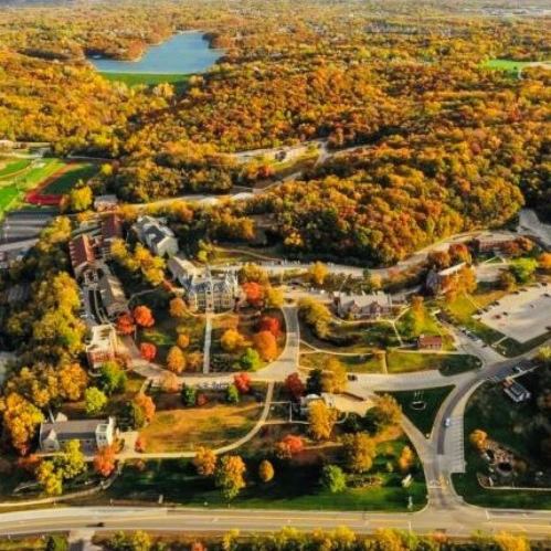 Aerial view of a colorful autumn landscape with trees, buildings, and winding roads near a lake.