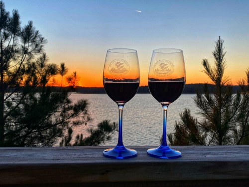 Two wine glasses filled with red wine, set against a vibrant sunset over a lake, framed by trees.