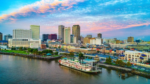 Aerial view of a city skyline at sunset, featuring a riverboat docked along the waterfront and colorful clouds above.