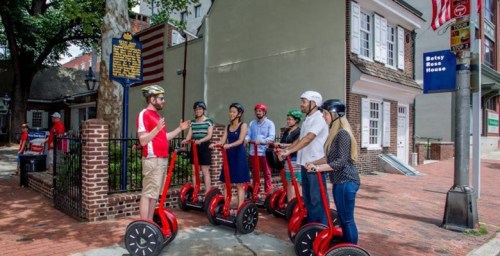 A group of people in helmets gathers around a guide near scooters in a historic area with buildings and an American flag.