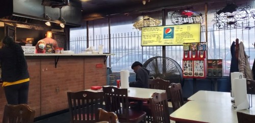 Interior of a casual restaurant with tables, a counter, and a menu board. A fan and a customer are visible.