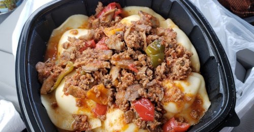 A takeout container filled with mashed potatoes topped with seasoned ground meat, peppers, and a savory sauce.