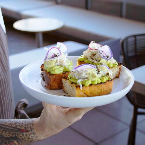 A hand holds a plate with three toasted bread slices topped with avocado, crab, and garnished with radish slices.