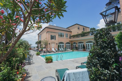 A sunny pool area surrounded by greenery and flowers, with lounge chairs and a building in the background.