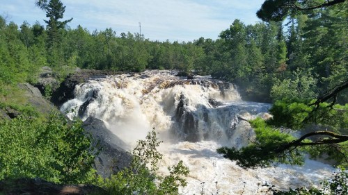 A powerful waterfall cascades over rocky cliffs, surrounded by lush green trees under a clear blue sky.
