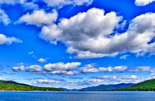 A serene lake surrounded by green hills under a bright blue sky filled with fluffy white clouds.