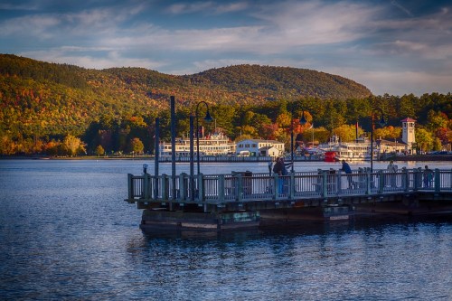 A scenic view of a pier by a lake, with boats and autumn foliage on the hills in the background.