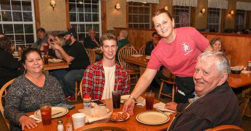 A waitress serves food to a smiling family at a cozy restaurant, with other diners visible in the background.