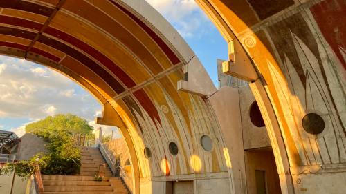 Architectural arches with colorful murals, bathed in warm sunlight, leading to a staircase and greenery in the background.